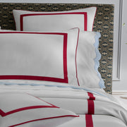 Close-up of white pillow shams bordered in red, a light blue sheet with a scalloped edge, dark patterned upholstered headboard.