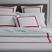 Bed with white pillow shams bordered in red, a light blue sheet with a scalloped edge, dark patterned upholstered headboard.