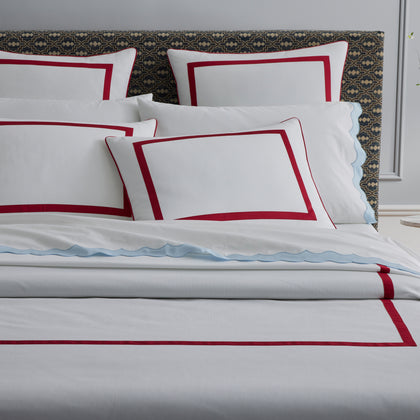 Bed with white pillow shams bordered in red, a light blue sheet with a scalloped edge, dark patterned upholstered headboard.