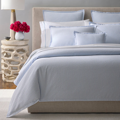 Pale blue-striped Matteo bedding styled on an upholstered bed, with pink peonies on a carved stone side table.