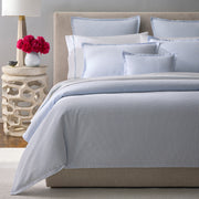 Pale blue-striped Matteo bedding styled on an upholstered bed, with pink peonies on a carved stone side table.