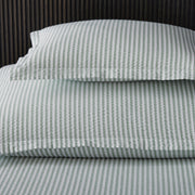 Close-up of sage green-striped Matteo pillow and sheets against a dark slatted wood headboard.