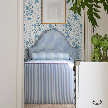 Pale blue-striped Matteo bedding on an upholstered headboard and bed, in an alcove with floral wallpaper and green ceramic lamps.