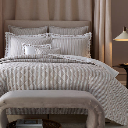 Taupe-striped Matteo quilted bedding set styled on a bed, with a curved bench and a wall-mounted lamp beside it.