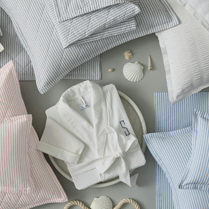 Overhead view of striped Matteo bedding in gray, pink, and blue, with a monogrammed white robe and seashells, from a different angle.