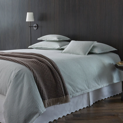 Sage green-striped Matteo bedding with a scalloped white bedskirt, styled on a bed with a brown fringed knit throw, against a dark striped wall.