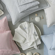 Overhead view of striped Matteo bedding in gray, pink, and blue, with a monogrammed white robe, seashells, and rope, arranged flat.