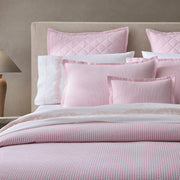 Pink-striped Matteo quilted bedding and pillows, styled on an upholstered headboard with embroidered white sheets.
