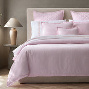 Pink-striped Matteo bedding styled on a bed with an upholstered headboard, quilted pillows, and embroidered white sheets.