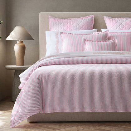 Pink-striped Matteo bedding styled on a bed with an upholstered headboard, quilted pillows, and embroidered white sheets.