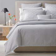 Gray-striped Matteo bedding styled on a bed with an upholstered headboard, quilted and seersucker pillows, with tulips on a nightstand.