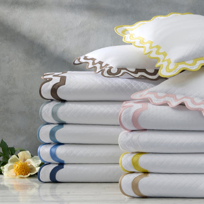 White Mirasol Matelasse quilted bedding with wavy trim in assorted colors, stacked beside a white flower.