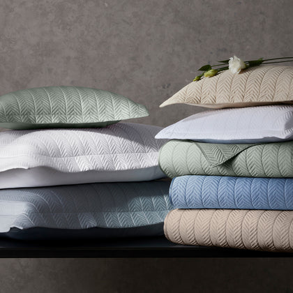 Netto chevron-textured quilted bedding stacked in sage, white, gray, and tan.
