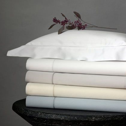 Nocturne Hemstitch sheets stacked in white, gray, ivory, and pale blue, on a dark table.