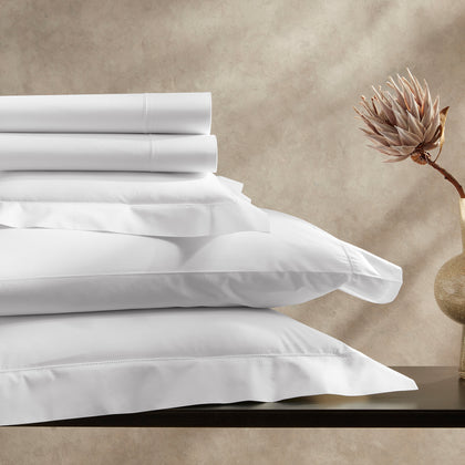 White Positano Hemstitch sheets stacked on a dark table, beside a dried protea flower.