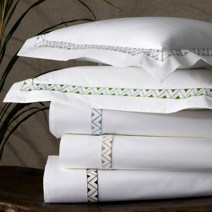 White Prado sheets with zigzag embroidered trim in assorted colors, stacked on a wooden table.