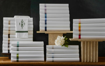 Stacks of white folded towels and linens with narrow colored stripes and a monogram logo, arranged on wooden display stands against a dark background.