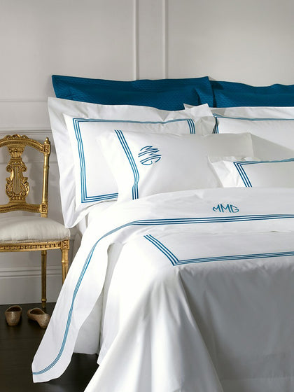White monogrammed bedding set with blue piping and monogrammed pillows, styled on a bed next to a gold chair.