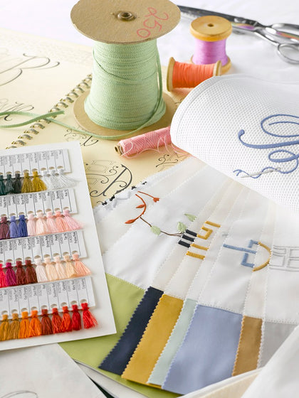 A styled flat-lay of embroidery supplies, including a spool of pale green thread, a card of thread color samples, white fabric swatches with embroidered lettering, and scissors on a light surface.