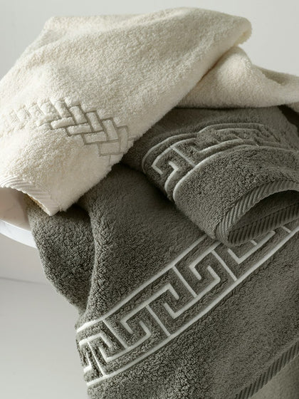 Two folded cotton towels, one cream and one taupe gray, both featuring an embroidered Greek key border, arranged on a white surface.