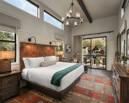 A rustic, modern bedroom featuring a bed with white linens and a green throw, set against a large copper-brown headboard. The room has white shiplap walls, a dark wood ceiling, a multi-light chandelier, and a red patterned rug.