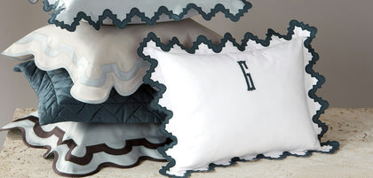A stack of decorative pillows with ruffled edges in white, navy, and gray, with a white front pillow featuring a dark navy monogram letter 'H'.