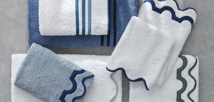 A coordinated set of white and blue towels with wavy trim, arranged on a light gray surface. The set includes bath towels, hand towels, and a washcloth, with some folded towels showing navy blue stripes and scalloped edges.