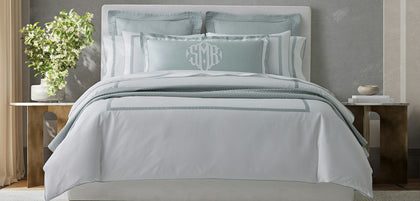 A neatly made bed dressed in white bedding with light blue-gray striped and solid accent pillows, including a monogrammed pillow, set in a styled bedroom with wooden bedside tables and a gray wall.