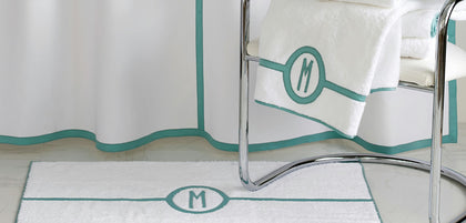 White bath mat and matching shower curtain with teal monogrammed letter 'M' and piping, shown in a bathroom setting with a chrome chair and white towel.