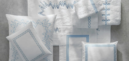 A coordinated set of white home textiles with blue embroidered geometric and zigzag patterns, including pillows, a folded towel, and a hanging cloth, arranged on a light gray surface.