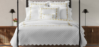 A neatly made bed with a white quilted coverlet, matching white pillows, and several white shams decorated with gold floral borders, set in a bedroom with dark wood nightstands and black table lamps.