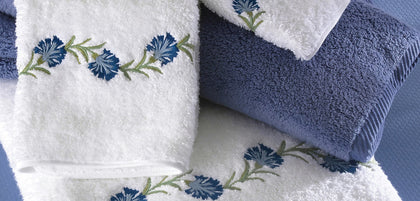 A set of folded towels and handkerchiefs in white and dusty blue, decorated with embroidered blue flowers and green stems.