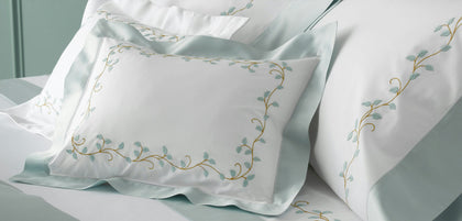 White satin pillowcases with light blue borders and gold embroidered vine detailing, styled on a bed.