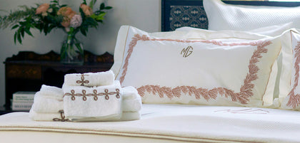A luxury bedding set featuring a cream-colored bedspread, a matching pillow with rose gold embroidered border and monogram, and folded white towels with rose gold embroidered details on a bed in a styled bedroom.