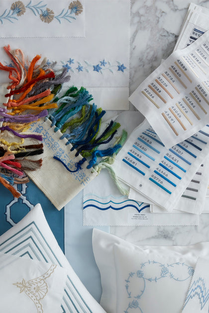 A styled flat-lay of embroidery supplies and finished embroidered fabric pieces on a marble surface. Visible items include multicolored embroidery floss/threads, labeled white paper swatches, embroidered white fabric panels, and a few finished decorative pillows or textile pieces with stitched motifs.