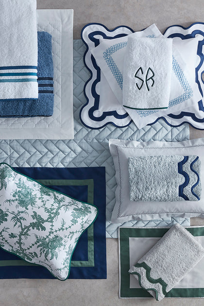 A coordinated bedding and bath set arranged in a flat lay on a light stone surface, including white towels with blue stripes, a blue towel, white pillows and sheets with blue scalloped trim, a white pillow with green toile-style border, and matching folded textiles.