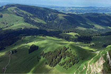 Aerial view of green alpine valleys and forested hills with a lake in the distance.