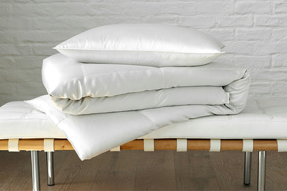 Edelweiss eiderdown pillow and comforter with a silky sheen, stacked on a bench.