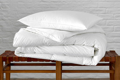 Valletto down pillow and rolled comforter stacked on a woven leather bench.