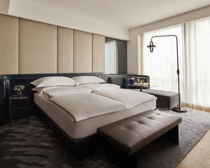 A modern luxury bedroom featuring a double bed with crisp white bedding, a tufted gray bench at the foot, a dark leather headboard, and a large patterned rug in a bright, contemporary room.