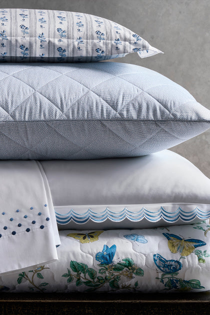 A stack of white and light blue bedding, including a quilted pillow, a folded sheet with blue embroidered dots, a pillowcase with a blue scalloped border, and a pillow with blue and yellow butterfly and floral prints.