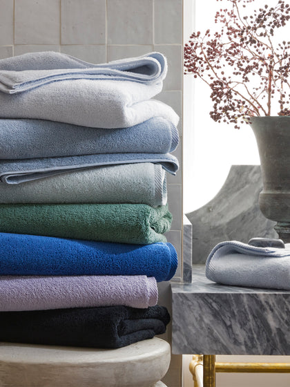 Milagro towels stacked in blue, sage, navy, and black tones on a marble pedestal