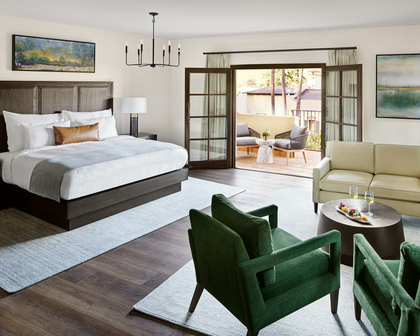 A luxury modern hotel suite featuring a large bed with white linens, a light beige sofa, two green velvet armchairs, dark wood floors, and large glass doors opening to a balcony.