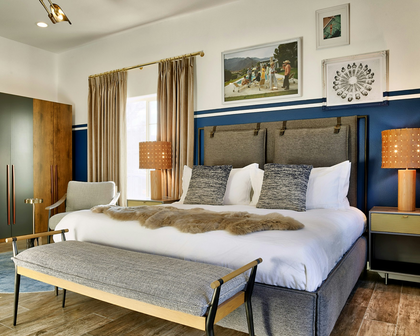 A styled bedroom featuring a bed with white linens, gray upholstered headboard and footboard, patterned gray pillows, a fur throw, a bench, curtains, lamps, and framed wall art against a blue-and-white wall.