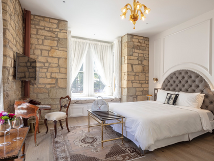 A luxury bedroom featuring a large bed with white linens and a tufted gray headboard, set in a room with rustic stone columns, a gold chandelier, and a patterned rug.