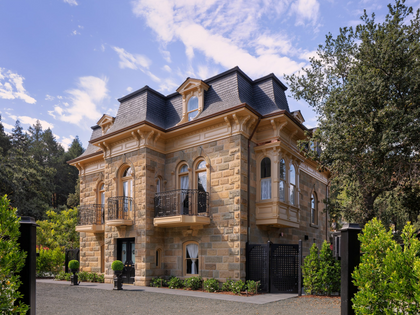 A grand stone villa with a dark slate roof, arched windows, and balconies, surrounded by lush green trees and a gravel driveway under a blue sky.