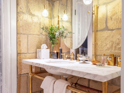 A luxury bathroom vanity setup featuring a white marble countertop and sink with a polished gold brass faucet and matching gold metal legs. The vanity is styled with a marble tissue box cover, a glass vase of greenery, and various toiletries, set against a rustic stone wall with a large oval mirror and hanging globe lights.