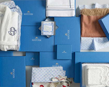 Flat lay of signature blue gift boxes with a monogrammed robe, folded sheeting, and a cashmere throw.