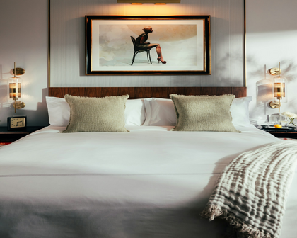 A neatly made bed in a modern bedroom setting, featuring white linens, two textured beige pillows, a dark wood headboard, bedside tables with lamps, and a framed art print on the wall above the bed.