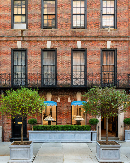 A brick building facade with black wrought-iron balconies, tall windows, and two storefront windows at the ground level. Two leafy trees in large light gray planters sit on either side of the entrance, with a paved walkway in front.